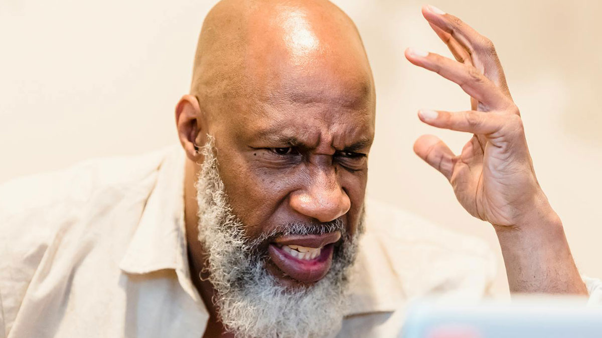 Man with gray beard showing frustration and confusion during a house ride conversation with his brother indoors.