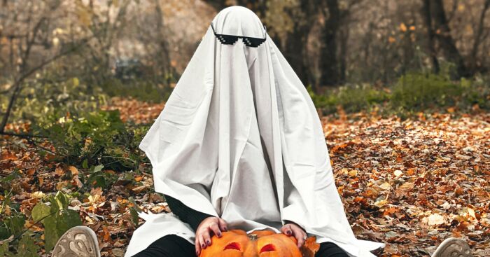 Person wearing a Halloween costume as a ghost with pixelated sunglasses sitting outdoors holding carved pumpkins.