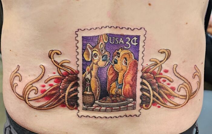Tattoo of a USA postage stamp featuring Lady and the Tramp with spaghetti, surrounded by decorative flames on lower back.