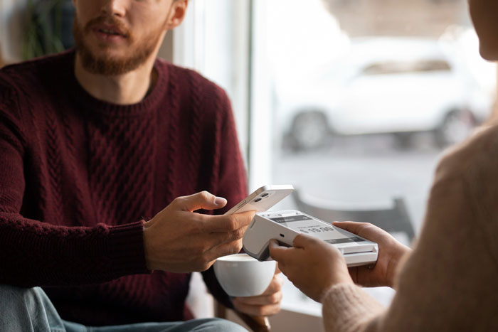 Man in a maroon sweater making a contactless payment with a smartphone while holding a coffee cup indoors. Man in a maroon sweater making a contactless payment with a smartphone while holding a coffee cup indoors.