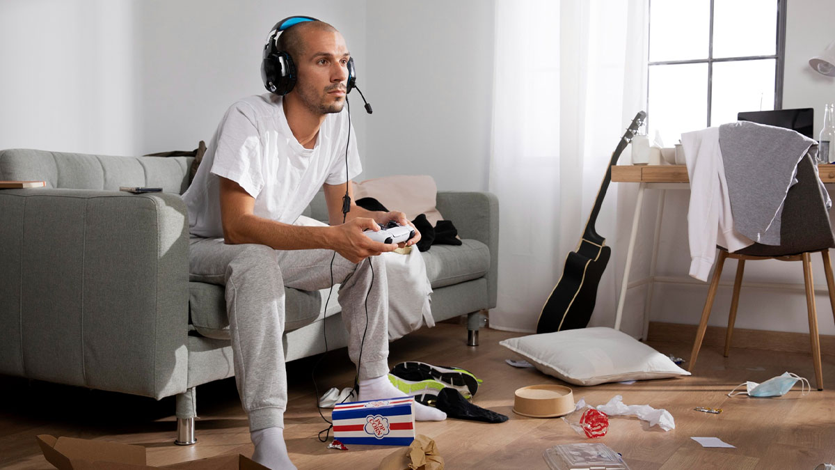 Young man wearing headphones, playing video games on a couch in a messy room, showing a friend helped with a place to stay.