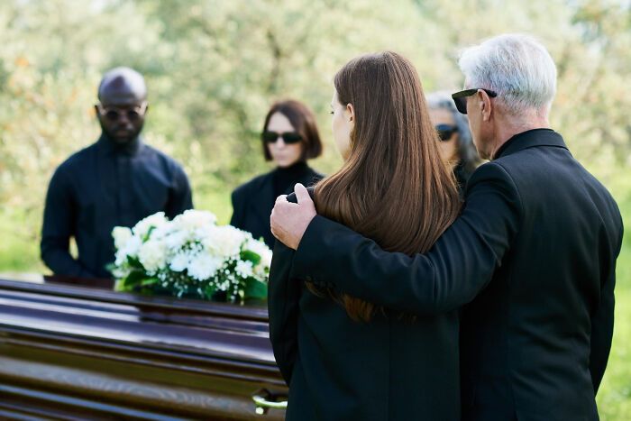 Mourning family at funeral, mother comforted by partner and ex-husband standing near a casket with flowers. Mourning family at funeral, mother comforted by partner and ex-husband standing near a casket with flowers.