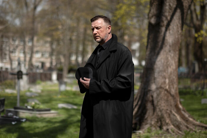 Man in black coat holding hat solemnly at a funeral, reflecting on mother, partner, and ex-husband relationships. Man in black coat holding hat solemnly at a funeral, reflecting on mother, partner, and ex-husband relationships.