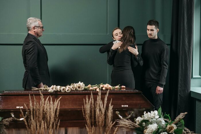 Mourners including mother, partner, and ex-husband comforting each other during a funeral beside a closed casket. Mourners including mother, partner, and ex-husband comforting each other during a funeral beside a closed casket.