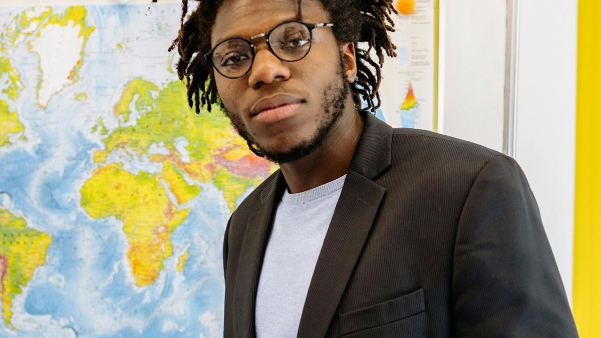 Young man wearing glasses and a black jacket standing in front of a world map, illustrating moments of embarrassment hilariously.