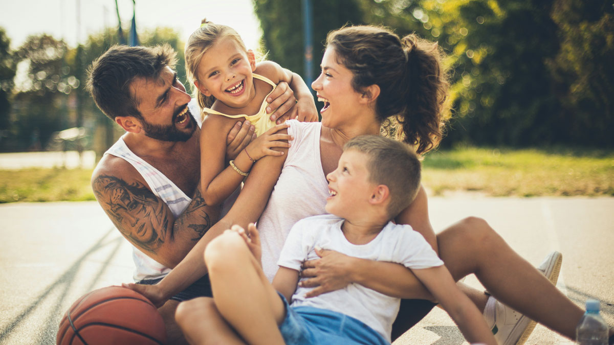 Happy family outdoors showing funny differences between moms and dads while playing basketball with children on a sunny day