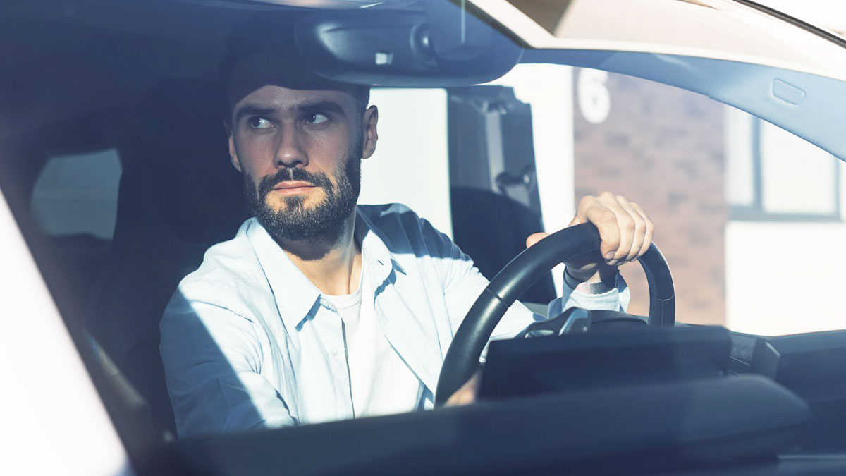 Bearded man wearing a light shirt sitting in a car, holding the steering wheel and looking intensely outside.