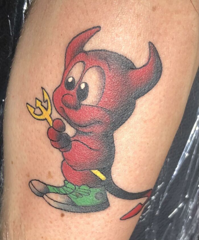 Tattoo of a cartoon-style red devil holding a yellow trident, wearing green shoes on a person's arm by tattoo artists.