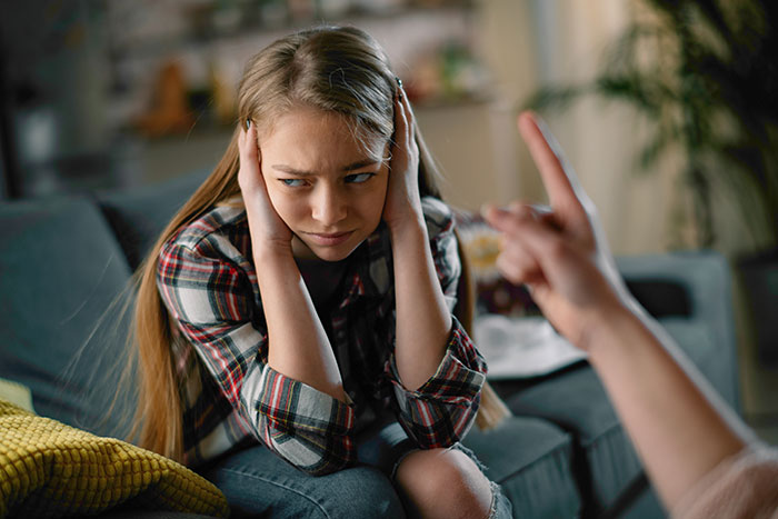 Teen girl sitting on a couch upset and covering ears while being scolded, showing anger and hurt feelings years later. Teen girl sitting on a couch upset and covering ears while being scolded, showing anger and hurt feelings years later.