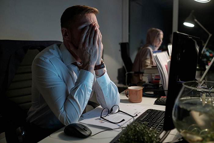 Man in office wearing white shirt, stressed and covering face, dealing with younger coworker flirt late at night.
