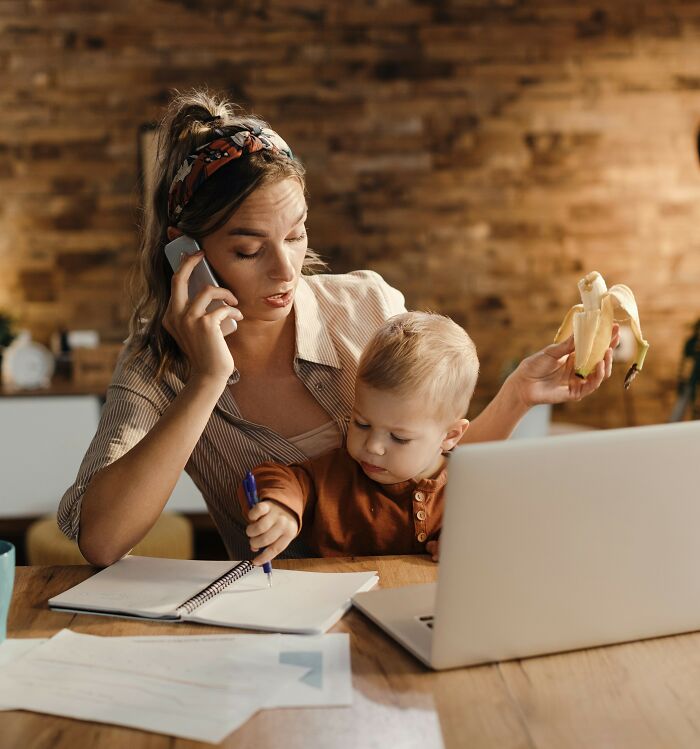 Female professor multitasking at home with child, eating banana, working on laptop, and talking on phone discussing evidence-based decision. Female professor multitasking at home with child, eating banana, working on laptop, and talking on phone discussing evidence-based decision.
