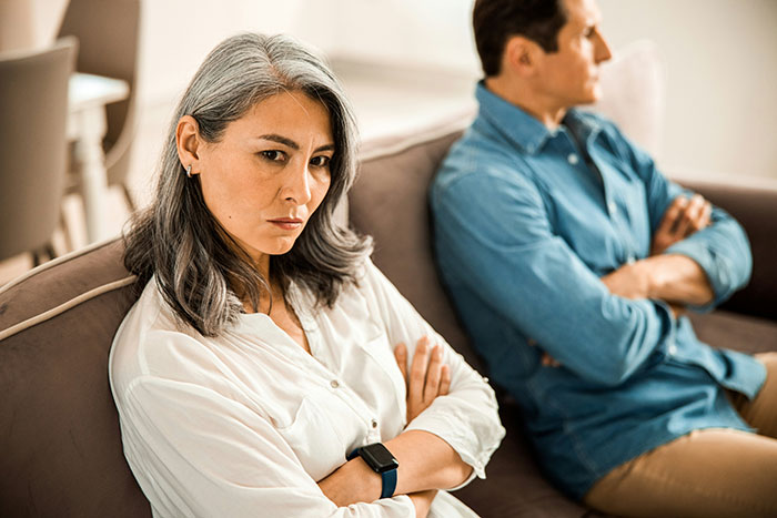 Middle-aged couple sitting apart on couch, showing tension and regret in an open marriage situation. Middle-aged couple sitting apart on couch, showing tension and regret in an open marriage situation.
