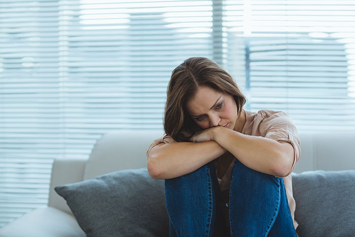 Sad woman sitting on couch hugging knees, showing signs of depression and emotional struggle in a bright room. Sad woman sitting on couch hugging knees, showing signs of depression and emotional struggle in a bright room.