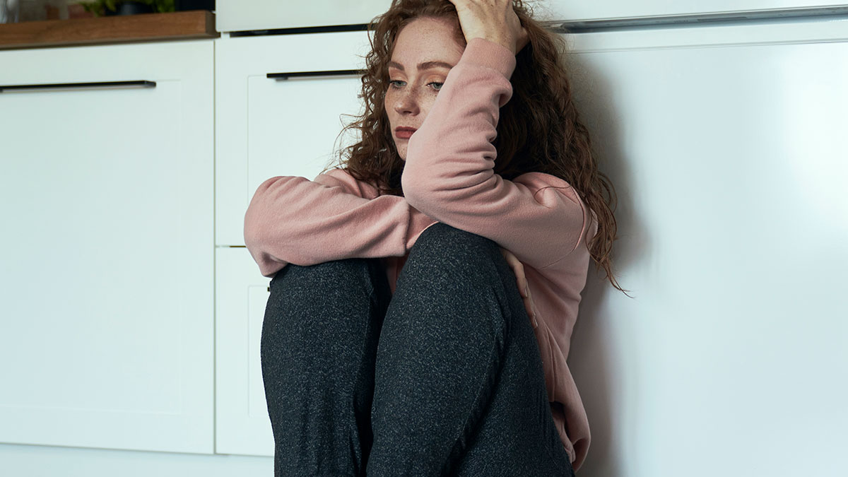 Young woman sitting on floor looking upset, illustrating depression and emotional struggle in a home setting.
