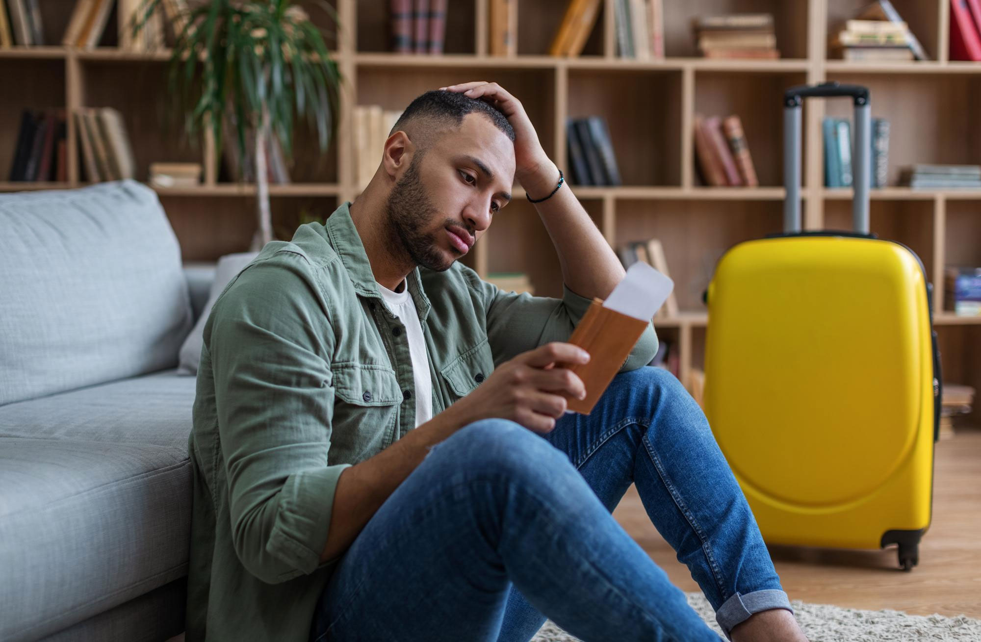 Man reconsidering relationship while holding travel document near yellow suitcase in living room with bookshelves. Man reconsidering relationship while holding travel document near yellow suitcase in living room with bookshelves.