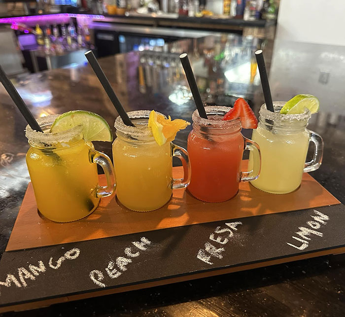 Four colorful fruit drinks in small glass jars with straws on a wooden tray at a restaurant bar setting.