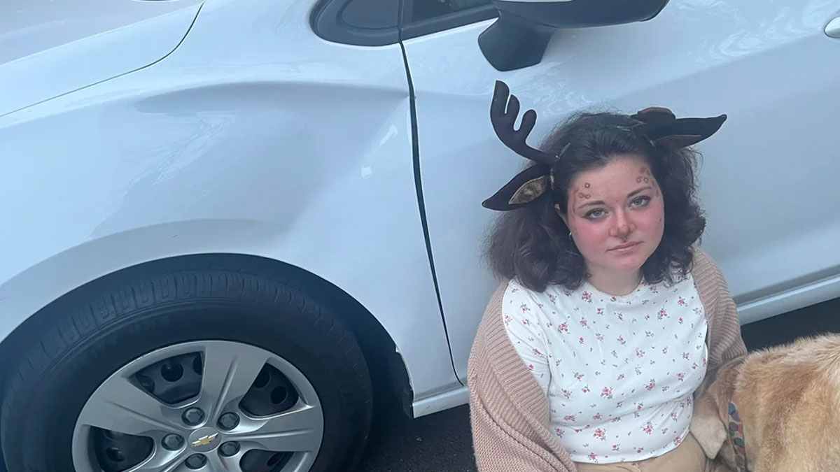 Young woman with deer antler headband posing next to a dented white car, showing a Halloween fail moment.