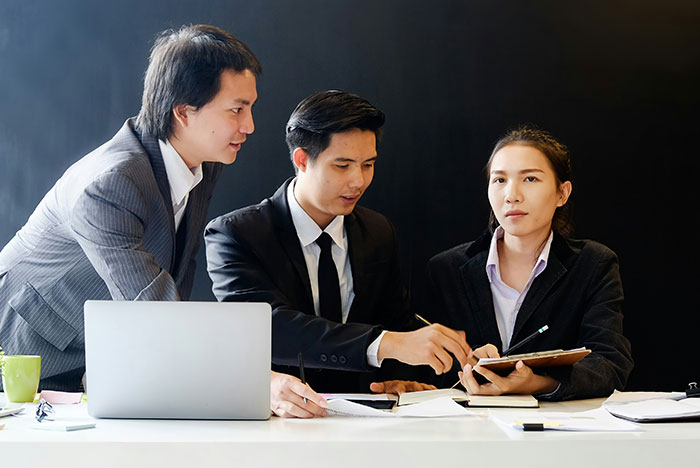 Three business professionals in formal attire discussing hard truths women realize while working together at a desk with a laptop.
