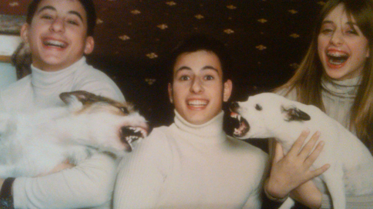 Three family members in white turtlenecks laughing as their dogs bark aggressively during a cringed family photo moment.