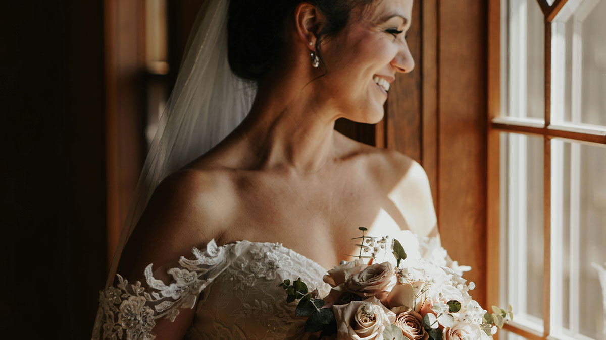 Bride in a lace off-shoulder wedding dress smiling while holding a bouquet near a wooden window with sunlight.