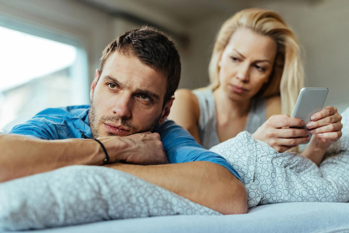 Couple on bed looking upset while woman holds phone, illustrating selfish SIL trying to ruin couple’s honeymoon conflict.
