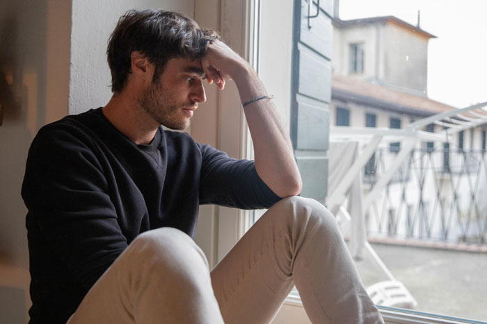 Young man sitting by window, looking heartbroken and distressed, reflecting emotional pain from past affair. Young man sitting by window, looking heartbroken and distressed, reflecting emotional pain from past affair.