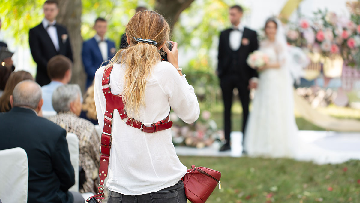 Woman wedding videographer capturing bride and groom at outdoor ceremony with guests seated in background