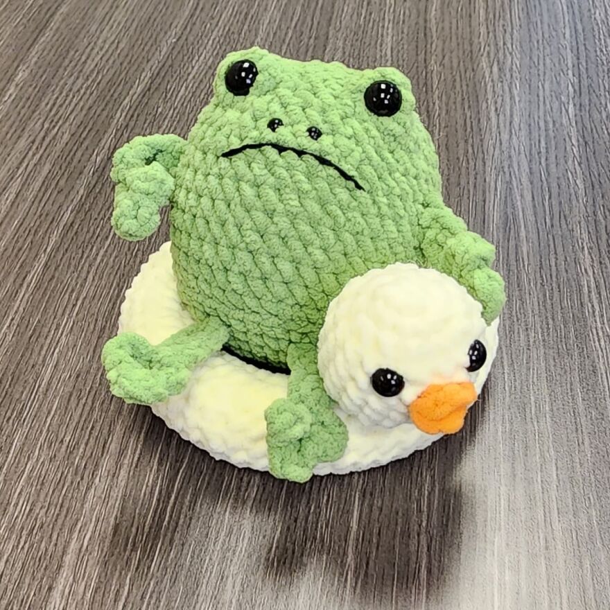 Crochet frog with big eyes sitting on a white crocheted duck, showcasing crochet animals with personality on wooden surface.