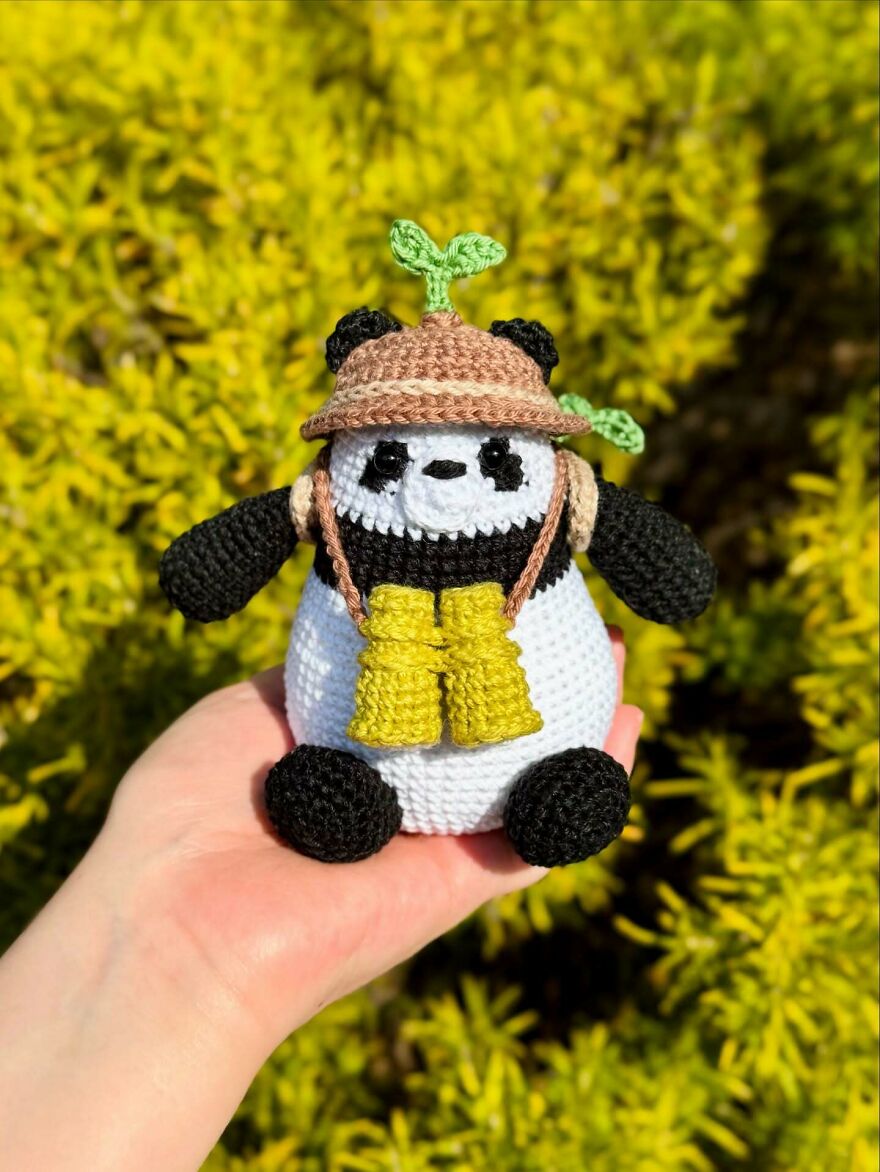 Crochet panda animal with binoculars and hat, held in hand against a bright yellow-green outdoor background.