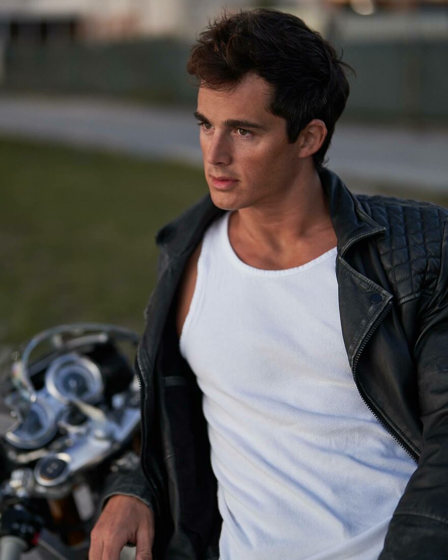 Young man wearing a leather jacket and white tank top, posing thoughtfully with a motorcycle, handsome man in the world 2025.
