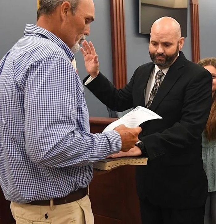 Police chief in black suit raising hand for oath while another man in checkered shirt reads a document indoors. Police chief in black suit raising hand for oath while another man in checkered shirt reads a document indoors.