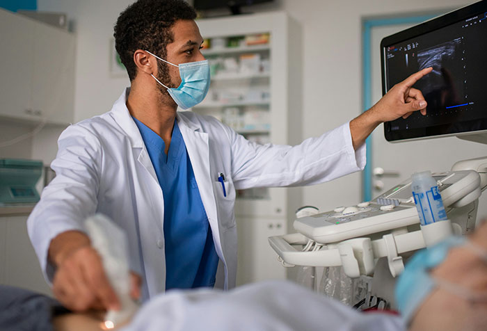 Doctor wearing mask and white coat performing ultrasound while reviewing self-diagnosed medical case on screen