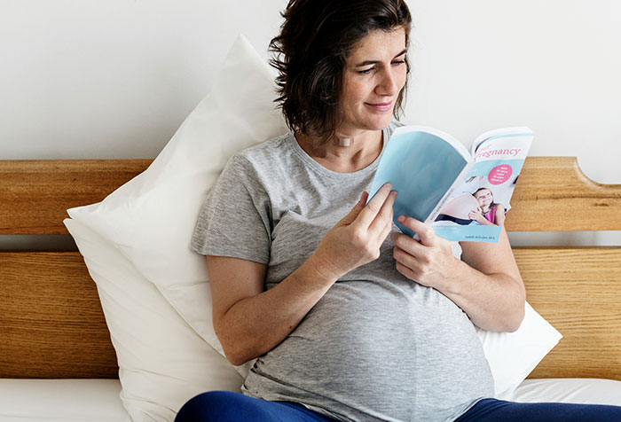 Pregnant woman sitting on bed reading a book about pregnancy, illustrating self-diagnosed health awareness.