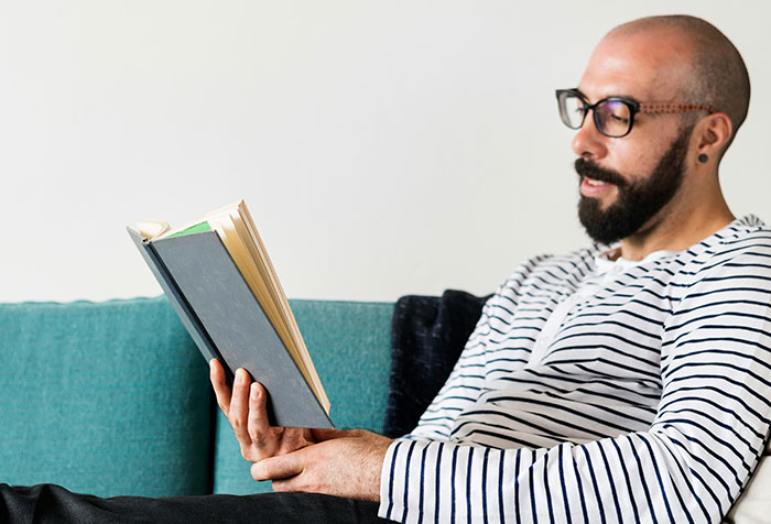 Man with glasses and beard reading a book on a couch, illustrating self-diagnosed health cases against initial doubt.