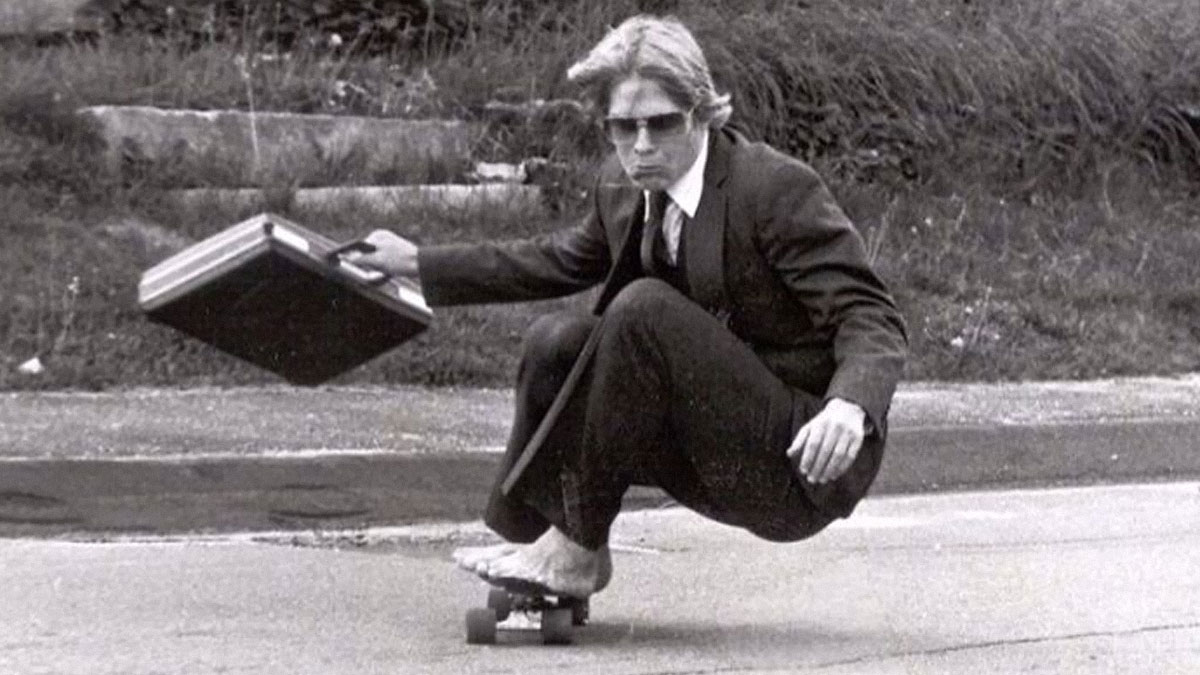Man in a suit riding a skateboard barefoot with a briefcase, a wild and weird vibe from the 1980s.