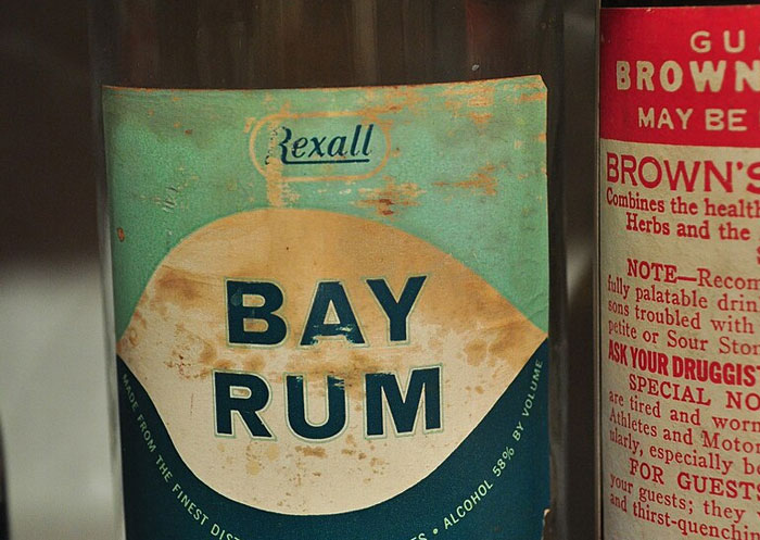 Close-up of a vintage Rexall Bay Rum bottle label with aged, faded graphics and text on a glass shelf.