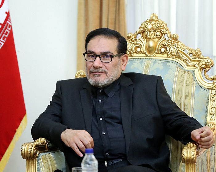 Iranian politician seated on an ornate chair with a red flag in the background amid risqué wedding dress outrage.