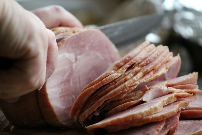 Hand slicing thin pieces of ham on a wooden board, related to shrimp jello and unusual guest foods.
