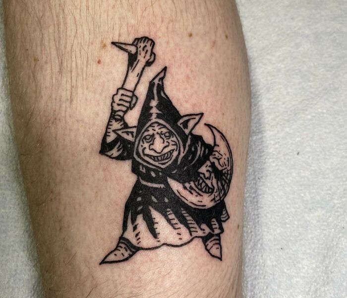 Tattoo of a goblin-like creature holding a crude weapon and a crescent moon on a person's forearm.