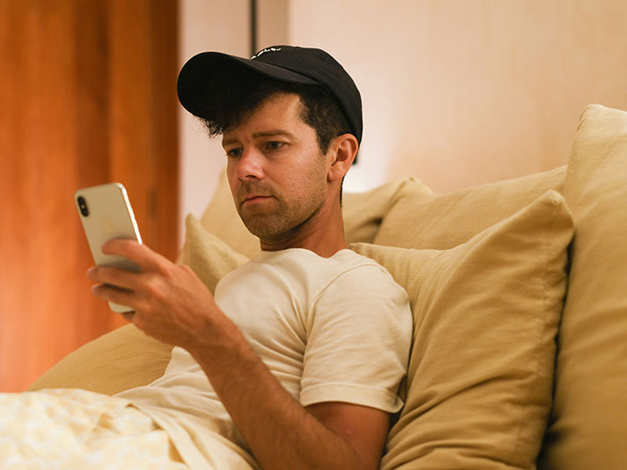 Man in a black cap sitting on a beige couch, looking serious while holding a smartphone during a family conflict.