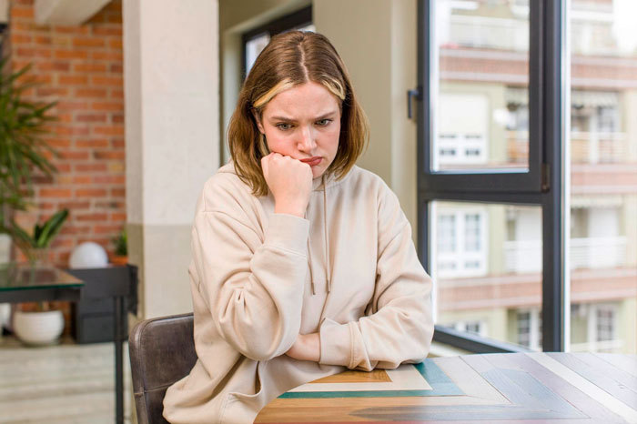 Young woman in a beige hoodie looking upset while sitting at a table, reflecting feelings of frustration over mooching friend. Young woman in a beige hoodie looking upset while sitting at a table, reflecting feelings of frustration over mooching friend.