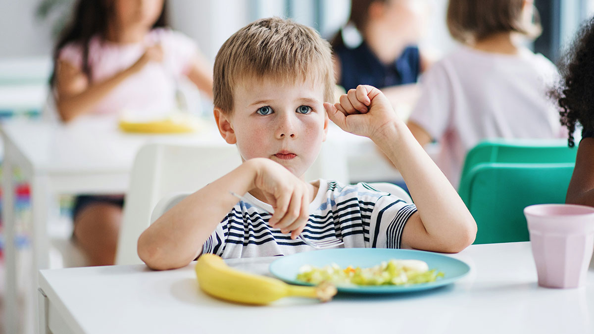 Young boy left hungry at school sitting with uneaten food and a banana on his plate during snack time.