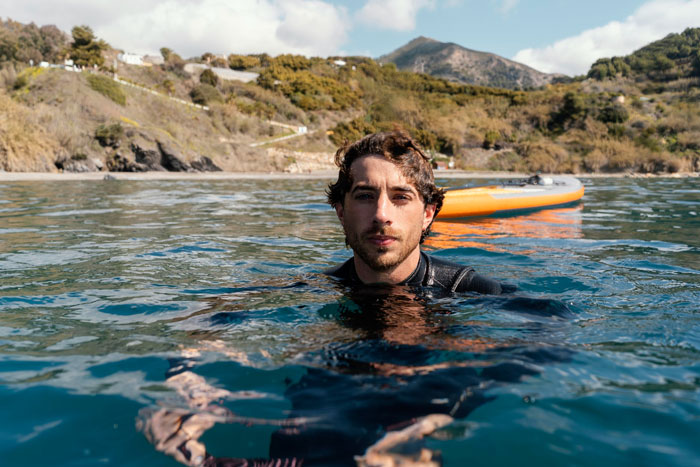 Man in wetsuit floating in the sea near a paddleboard with coastal hills in the background, elite military diver concept. Man in wetsuit floating in the sea near a paddleboard with coastal hills in the background, elite military diver concept.