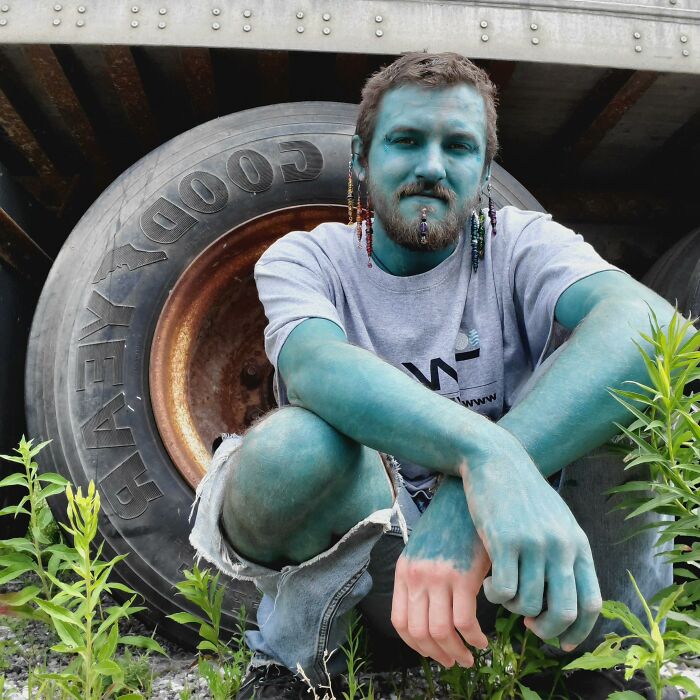 Man with full blue body paint and colorful braided hair sitting in front of a large tire, related to strange tattoo artists topic.