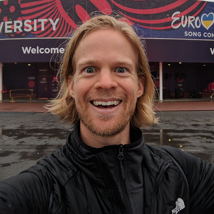 Man behind blinking guy GIF selfie with long hair and beard, smiling outside a building with colorful banners. Man behind blinking guy GIF selfie with long hair and beard, smiling outside a building with colorful banners.