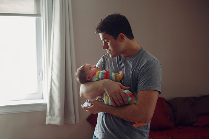 Father holding and looking at baby in a dimly lit room, illustrating burnt-out wife asking husband to feed the baby.