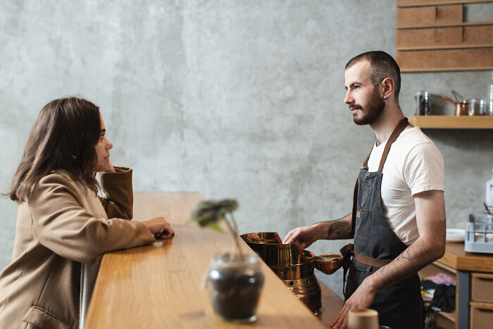 Waiter in apron talking to a single female customer at a wooden counter in a casual dining setting.