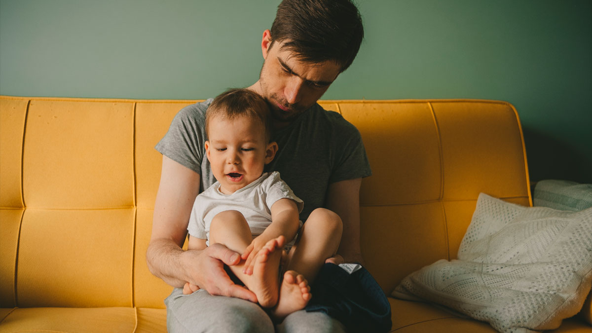 Man sitting on yellow couch holding young son who has no idea about his real parents in cozy home setting