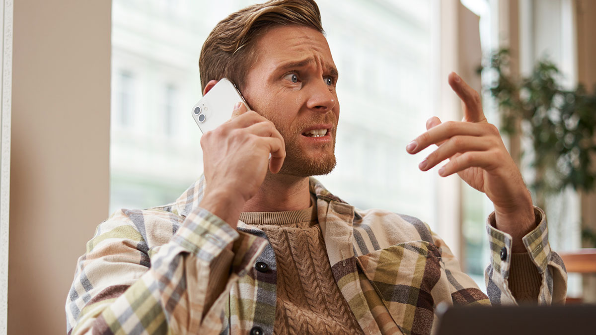 Man in plaid shirt talking on phone with frustrated expression inside a cafe, depicting rejection and dating situation.