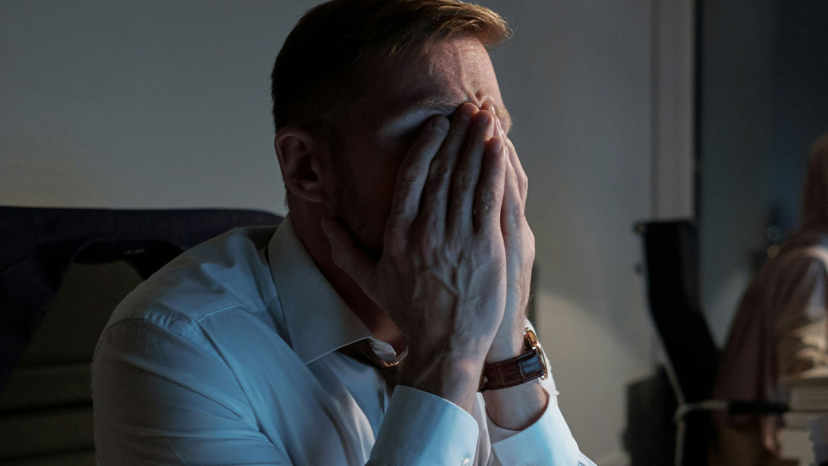 Stressed man in office covering face with hands, feeling overwhelmed after shutting down younger coworker's flirt.